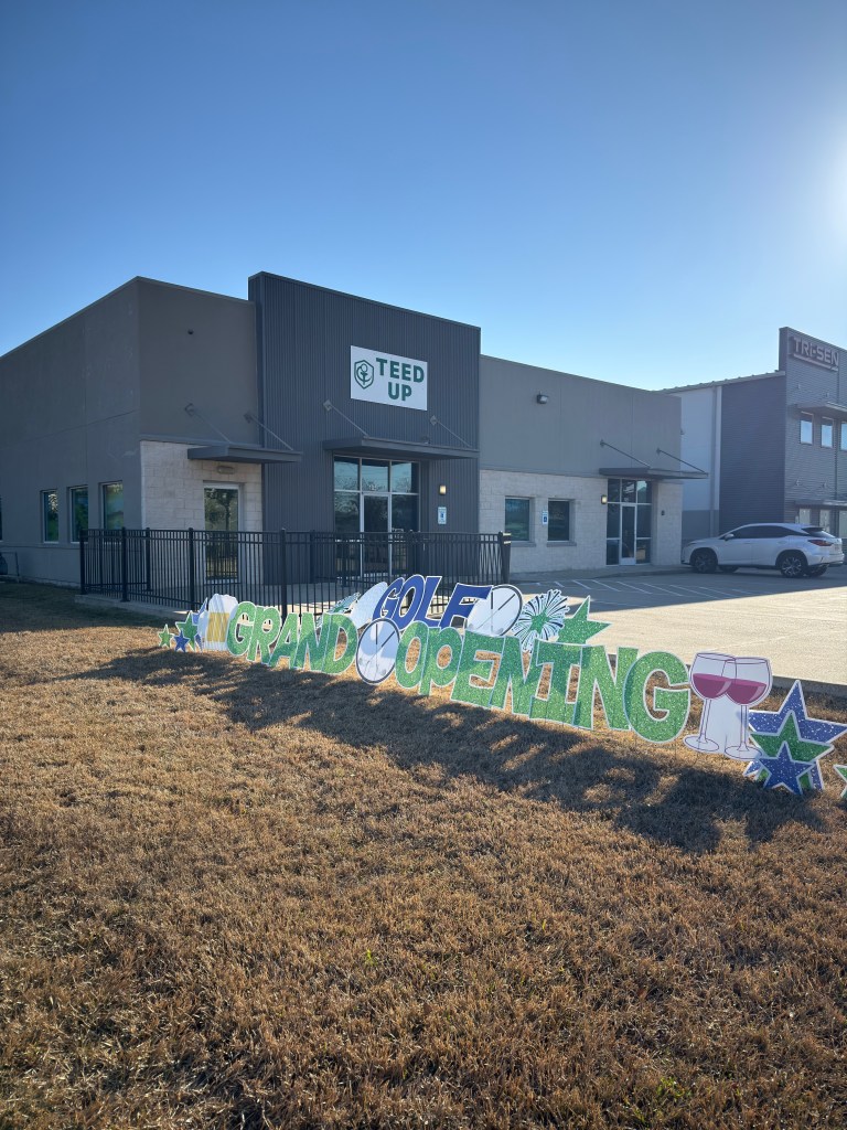 gray building in webster tx with white signage saying teed up on the awning and a large yard sign saying golf grand opening in front of the parking lot